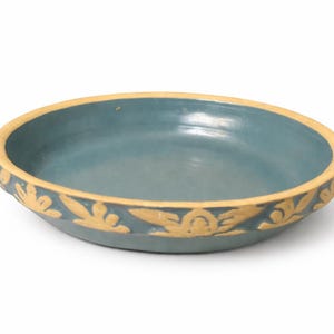 May include: A shallow, round, teal-colored ceramic bowl with a decorative border. The rim is a light tan color, and the border features a repeating gold leaf design. The bowl is set against a white background.