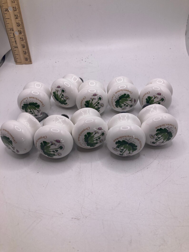 May include: Ten white ceramic cabinet knobs with a floral design. Each knob features a green and white illustration of a Chrysanthemum flower with the text "Chrysanthemum" written around the flower.