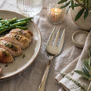 May include: A plated meal featuring sliced chicken breast, roasted potatoes, and green beans. A silver fork rests on the linen tablecloth, next to a lit candle in a glass holder, a small bowl of salt, and a decorative vase with greenery.