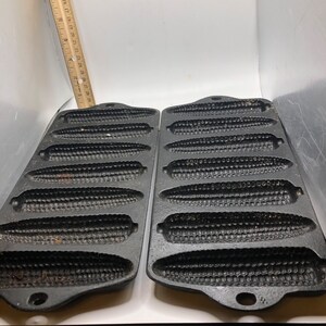 May include: Two black cast iron cornbread pans with six individual molds shaped like corn cobs. The pans have a handle on each side.