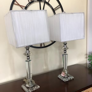 May include: Two elegant table lamps with square white shades. The lamps have a clear, faceted crystal base and a silver-toned metal accent. The lamps are placed on a dark brown wooden surface, with a large black clock in the background.