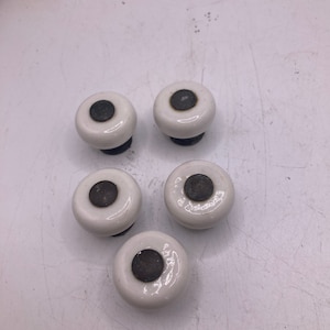 May include: Five white ceramic drawer knobs with black centers. The knobs are round and have a smooth, glossy finish.