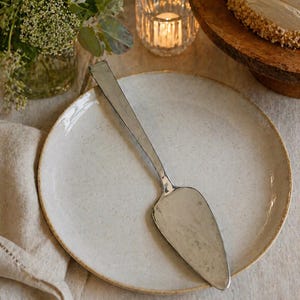 May include: A white ceramic plate with a silver cake server. The plate has a slightly textured surface and a light brown rim. The server is resting on the plate, ready to serve a cake. The scene is set on a beige linen tablecloth.