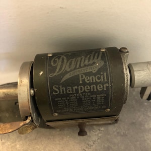 Op de afbeelding: Een antieke Dandy Automatic Feed Pencil Sharpener. De metalen puntenslijper is donkergroen met de woorden "Dandy Automatic Feed Pencil Sharpener" op de voorkant. De puntenslijper is gepatenteerd onder verschillende data.
