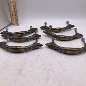 May include: Six antique brass cabinet door pulls with a decorative design. The pulls have a curved shape and a pointed end. The pulls are in good condition with some signs of wear.