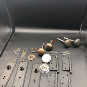 May include: An assortment of antique door hardware, including doorknobs, hinges, and backplates. The doorknobs are in various styles, with finishes ranging from brass to dark metal. The hinges are black, and the backplates have keyholes.