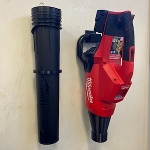 May include: A red and black cordless leaf blower with a black nozzle and a black storage tube hanging on a white wall. The leaf blower has the text "Milwaukee" and "FUEL" on it.