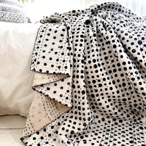 Pode incluir: Um cobertor creme com um padrão repetido de bolinhas pretas. O cobertor tem uma borda ondulada com costura preta. O cobertor é drapeado sobre uma superfície branca, criando uma estética aconchegante e convidativa. O cobertor é feito de um material macio.