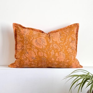 May include: A rectangular cushion with a floral pattern in shades of orange and peach. The cushion has a stitched design and a ruffled edge. The cushion is placed on a white surface.