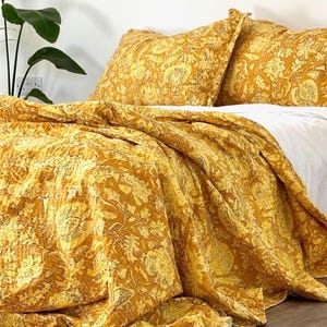 May include: A bed dressed with a mustard yellow quilt and matching pillowcases. The bedding displays a detailed floral pattern in shades of yellow and grey. The quilt is draped over a white sheet, creating a layered effect. A houseplant is visible in the background.