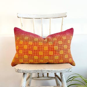 May include: A rectangular throw pillow featuring a vibrant gradient of pink, orange, and yellow hues. The fabric displays a repeating grid pattern of small squares with visible running stitch embroidery throughout the entire surface of the pillow.