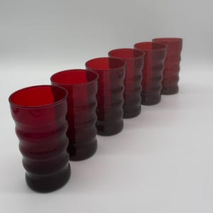 May include: A set of six red-tinted drinking glasses. Each glass has a unique, wavy, textured design. The glasses are arranged in a diagonal line, showcasing their shape and color against a white background. The glasses are made of glass.