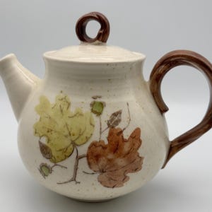 May include: A ceramic teapot with a cream-colored speckled glaze, featuring a brown twisted handle and lid knob. The body is decorated with painted illustrations of green and brown autumn leaves and small acorns on thin branches.
