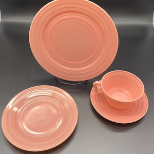 May include: A set of three pink ceramic dishes, including a large dinner plate, a smaller salad plate, and a teacup and saucer.