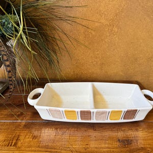May include: A rectangular, off-white ceramic serving dish with two compartments and rounded handles. The dish is decorated with a repeating pattern of brown, tan, and yellow rectangles. The dish is on a wooden surface.