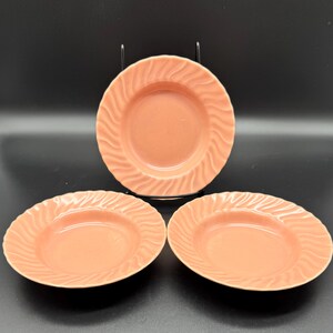 May include: Three peach-colored ceramic plates with a wavy, textured rim. The plates are arranged on a black surface, with one plate elevated on a metal stand. The plates are round and have a shallow depth, suitable for serving pasta or salads.