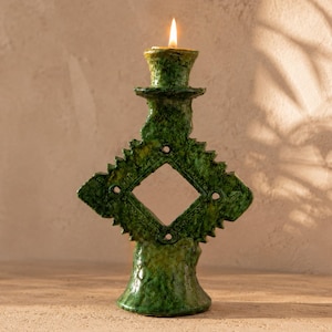 Puede incluir: Un candelabro de cerámica verde oscuro con textura y acabado brillante. La estructura presenta un recorte central en forma de diamante rodeado de bordes dentados. Una vela encendida se encuentra en la copa superior del objeto.