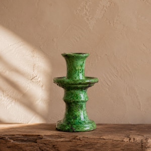 May include: A vibrant green ceramic candle holder with a glossy, mottled finish. The object features a wide, flared top opening, a central circular drip plate, and a sturdy, tiered pedestal base with a textured, uneven surface throughout.