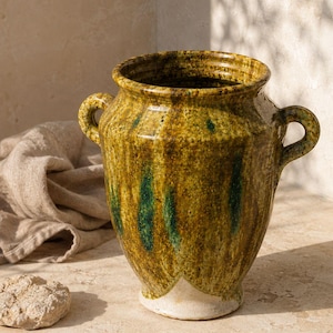 May include: A ceramic vessel featuring a mottled mustard yellow and deep green glaze with a glossy finish. The piece has a wide, circular opening, two small side handles, and a tapered base that reveals a light cream-coloured unglazed bottom.