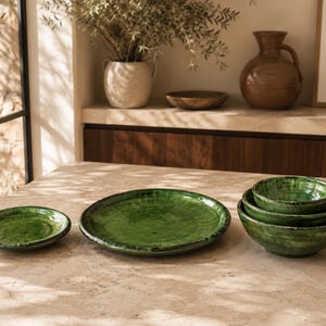Puede incluir: Un juego de vajilla de cerámica verde que incluye un plato pequeño, un plato grande y una pila de tres cuencos anidados. Cada pieza tiene un acabado brillante y moteado con variaciones de tonos verde bosque profundo y verde esmeralda.