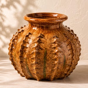 May include: A round, bulbous ceramic vase with a glossy, mottled brown glaze and a wide, circular opening. The exterior surface features prominent, vertical, serrated ridges that run from the base to the neck, accented by subtle green streaks.