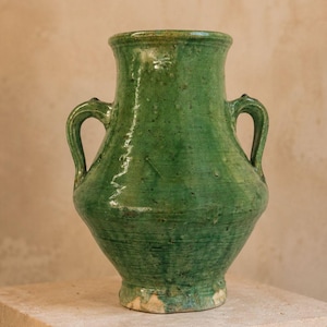 May include: A green glazed ceramic vase with a wide neck, a rounded body, and two symmetrical side handles. The surface features a glossy, mottled finish with visible texture, variations in color intensity, and a slightly uneven base edge.