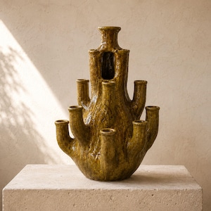 May include: A multi-spouted ceramic vase with a glossy, mottled olive-green glaze and a textured, bark-like surface. The vessel features a central vertical column surrounded by several smaller, protruding tubular openings for individual stems.