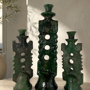 May include: A set of three dark green glazed ceramic candle holders with a textured, jagged edge design. Each holder features a central vertical column with three circular cutouts and a flared base, standing at varying heights in a group.