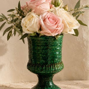 Könnte beinhalten: Eine tiefgrüne Keramikvase auf einem Sockel mit strukturierter, gesprenkelter Oberfläche, gefüllt mit einem Blumenstrauß aus hellrosa und cremefarbenen Rosen, akzentuiert durch grüne Eukalyptusblätter und kleine Zweige.