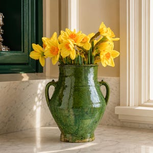 May include: A glossy, dark green ceramic vase with two side handles, filled with a vibrant bouquet of bright yellow daffodils. The vase features a rounded body and a flared rim, showcasing a smooth, reflective glaze and subtle texture.