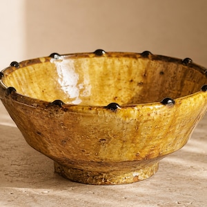 May include: A round, glazed ceramic bowl with a textured, mottled mustard-yellow finish. The rim features a series of small, evenly spaced, dark brown raised dots that encircle the top edge, providing a distinct tactile detail to the piece.