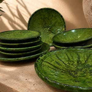 Op de afbeelding: Een verzameling ronde keramische borden met een levendig, glanzend groen glazuur en een getextureerd, organisch oppervlak. De set bevat een stapel kleinere borden en diverse grotere, ondiepe borden met onregelmatige, handgemaakte randen.
