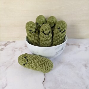May include: A white ceramic bowl filled with six green crocheted pickle toys with smiling faces. One pickle toy is sitting outside the bowl on a white marble surface.