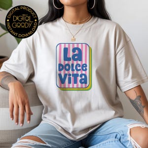 May include: A beige t-shirt featuring a colorful graphic that reads "La Dolce Vita" in blue and white. The graphic is set within a pink and white striped rectangle with a yellow and green border. The shirt is worn by a person.