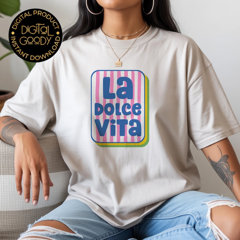 May include: A beige t-shirt featuring a colorful graphic that reads "La Dolce Vita" in blue and white. The graphic is set within a pink and white striped rectangle with a yellow and green border. The shirt is worn by a person.