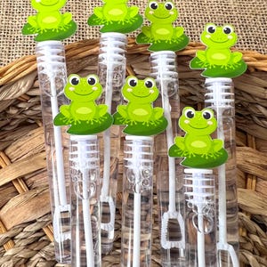 May include: Clear plastic bubble wands topped with green frog-shaped decorations. Each frog sits on a green lily pad. The wands have white handles and are arranged against a woven background.
