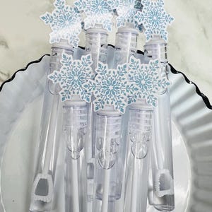 May include: Clear plastic bubble wands topped with light blue and white snowflake decorations. The wands are arranged in a white, scalloped-edge dish. The bubble wands are a festive item for winter events.