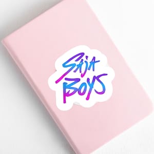 May include: A white sticker with the text "Saja Boys" in a gradient of blue and purple, placed on a pink notebook. The sticker has a handwritten style font.
