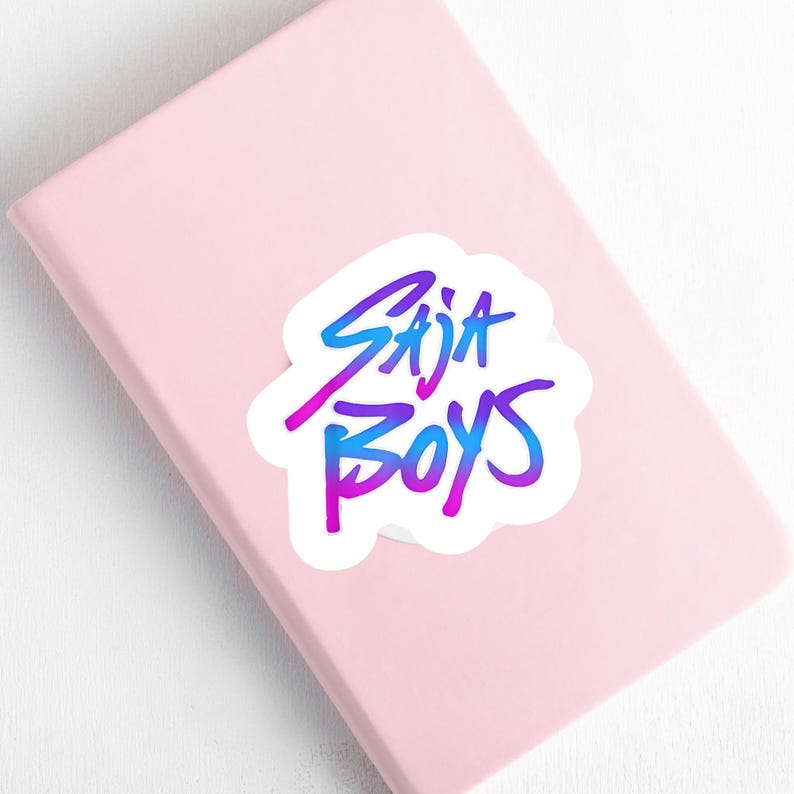 May include: A white sticker with the text "Saja Boys" in a gradient of blue and purple, placed on a pink notebook. The sticker has a handwritten style font.