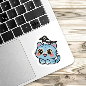 May include: A sticker featuring a cartoon blue cat with big yellow eyes and a black and white bird perched on its head. The cat has a happy expression and is sitting down. The sticker is on a laptop.