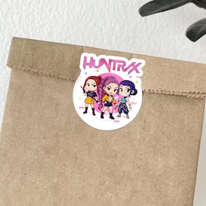 May include: A round sticker with the text "HUNTRAX" and three cartoon characters. The characters are in yellow, purple, and blue outfits. The sticker is on a brown paper bag.