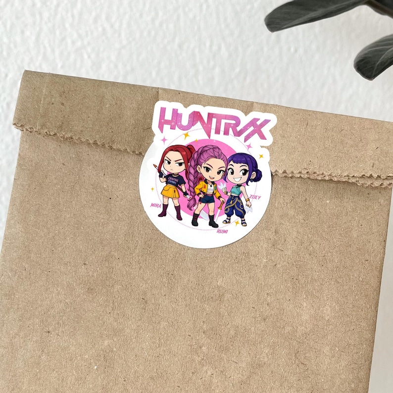 May include: A round sticker with the text "HUNTRAX" and three cartoon characters. The characters are in yellow, purple, and blue outfits. The sticker is on a brown paper bag.