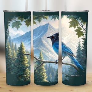 Può includere: Un bicchiere con una dettagliata illustrazione di un uccello blu posato su un ramo, con uno scenario di montagna e foresta. Il design è incorniciato da un bordo verde scuro con foglie d'acero. Il bicchiere ha una cannuccia bianca.