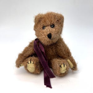 以下が含まれることがあります： 首に濃い紫色のリボンを巻いた、小さな茶色のテディベア。クマは黒い鼻と目をしており、足のパッドはクリーム色です。毛皮は柔らかく、質感のある素材で、クマは直立しています。