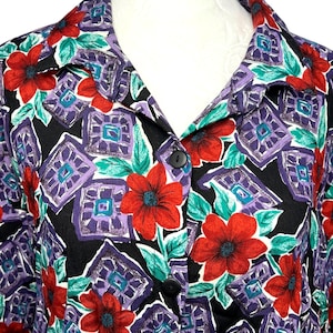 May include: A patterned button-down shirt with a floral design. The shirt displays large red flowers, teal leaves, and geometric shapes in purple and teal on a black backdrop. The collar and buttons are visible.