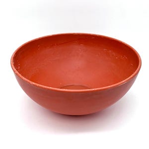 May include: A round, terracotta-colored bowl with a smooth interior and a slightly textured exterior. The bowl has a wide opening and a rounded base, suitable for serving or display. The bowl is made of a solid material and is set against a white background.