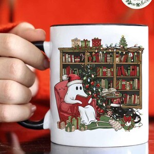 May include: White ceramic mug with a black handle featuring a Christmas-themed illustration of a ghost wearing a Santa hat reading a book in a red armchair by a decorated Christmas tree in front of a bookshelf filled with books. The ghost is sitting next to a black cat wearing a Santa hat.