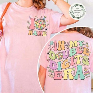 May include: A light pink t-shirt with a colorful retro design. The front features the number 10, a disco ball, and the word "NAME". The back says "IN MY DOUBLE DIGITS ERA" with additional graphics. The Ecoart Gallery logo is in the top right.