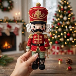 May include: A crocheted nutcracker doll featuring a red jacket with gold buttons, a tall red hat with gold embroidery, white hair curls, a black moustache, and black boots. The figure holds a small gold sceptre and wears green trousers.