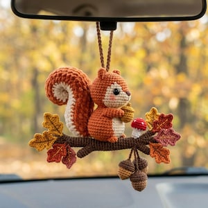 May include: A crocheted squirrel ornament hanging from a brown cord. The squirrel is orange and cream, holding a brown acorn. It sits on a brown branch decorated with colorful crocheted autumn leaves, a small red mushroom, and three hanging acorns.